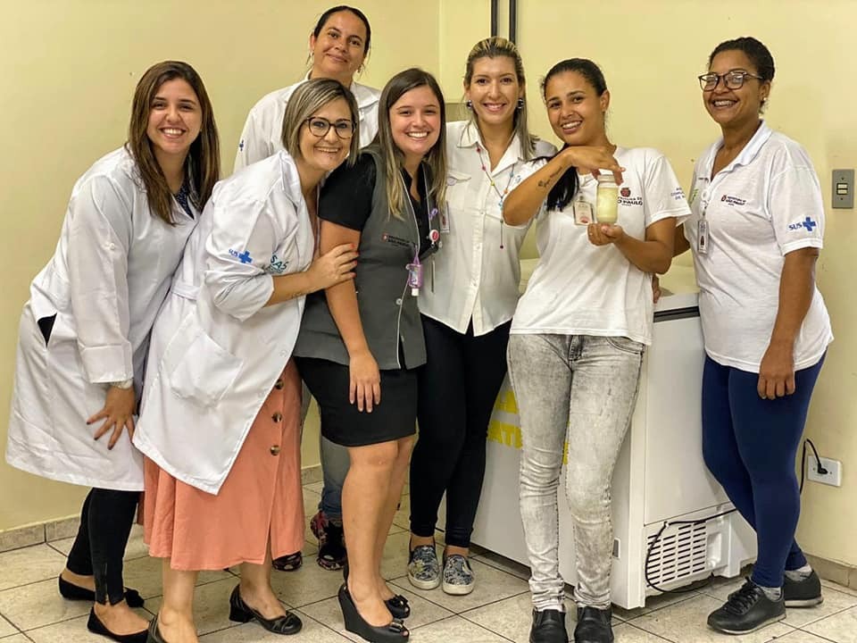 Na imagem há sete mulheres posando para a foto. Uma delas está segurando uma mamadeira com leite humano dentro. Todas sorriem para a foto.